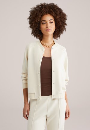 Mladá žena s kučeravými vlasmi, ktorá nosí zlatý reťaz, krémový kabát a nohavice a hnedé rebrované tielko, stojí pred béžovým pozadím.