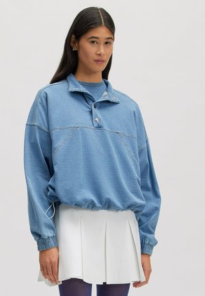 Vrouw met lang donker haar, gekleed in een lichtblauwe trui met drukknopen en een witte geplooide rok tegen een effen achtergrond.