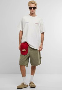 White oversized t-shirt with red embroidered text, olive green shorts, red baseball cap, and brown slip-on shoes with white socks.