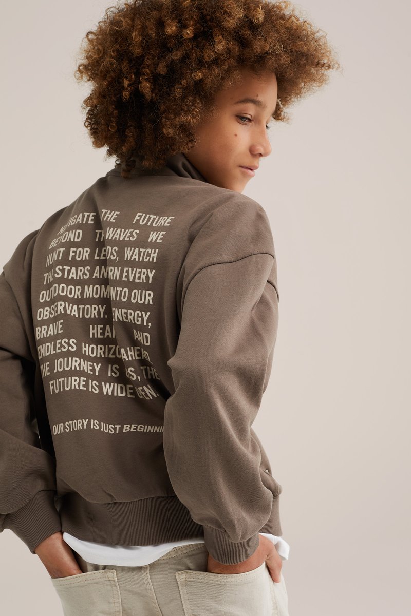 Jeune personne aux cheveux bouclés portant un sweat-shirt marron avec un texte blanc au dos, debout les mains dans les poches d'un pantalon clair.