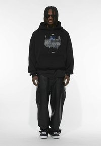 Sudadera negra con estampado gráfico, combinada con pantalones cargo de cuero negro y zapatillas negras. Diseño elegante con corte holgado y tejido texturizado.
