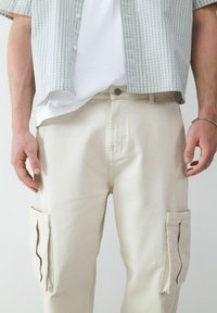 Beige cargobroek met zijzakken, knoopsluiting en een gladde textuur. Gecombineerd met een groen geruite shirt en een wit T-shirt.