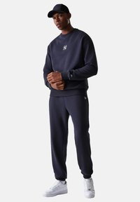 Joven con gorra negra, sudadera negra de NY, pantalones deportivos negros a juego y zapatillas blancas, de pie con las manos entrelazadas.