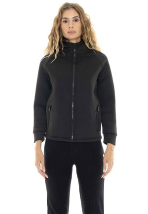 Une femme porte une veste noire zippée à col montant et dotée de deux poches zippées, avec un pantalon noir, sur fond blanc.