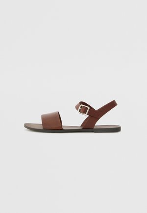 Sandale en cuir marron avec une large lanière avant, une lanière de cheville ajustable avec boucle et une semelle en caoutchouc plate. Design minimaliste et texture lisse.