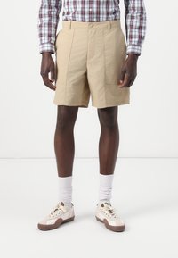 Beige shorts with a textured finish, featuring a waistband, pleated front, and side pockets. Paired with white socks and casual sneakers.