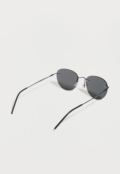 Λεπτά γυαλιά ηλίου με μαύρο μεταλλικό πλαίσιο, σκούρα γκρι οβάλ φακούς και ρυθμιζόμενα μαξιλαράκια μύτης, τοποθετημένα πάνω σε λευκή επιφάνεια.