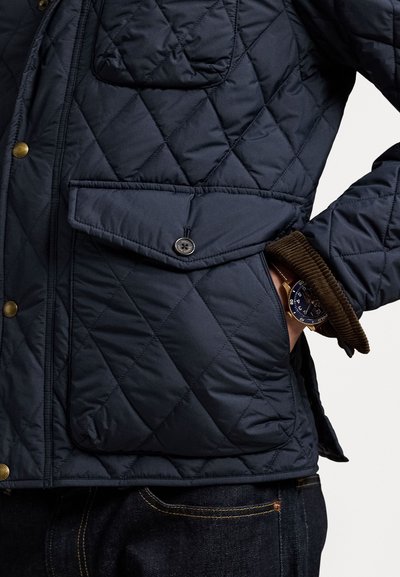 Gestepptes dunkelblaues Jacket mit diagonalen Nähten, ausgestattet mit einer Brusttasche und einer unteren Klapptasche. Sichtbares braunes Corduroy-Bündchen mit einer Uhr.
