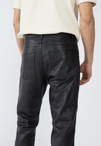 Schwarze Lederhosen, glatte Textur, schlanker Schnitt, zwei Gesäßtaschen, gestickte Details, getragen mit einem hellen Oberteil.