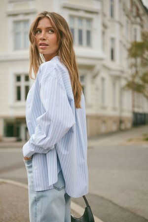 Frau mit langen Haaren, die ein hellblau gestreiftes Hemd und Jeans trägt, steht im Freien nahe einem Gebäude und hält eine schwarze Handtasche.