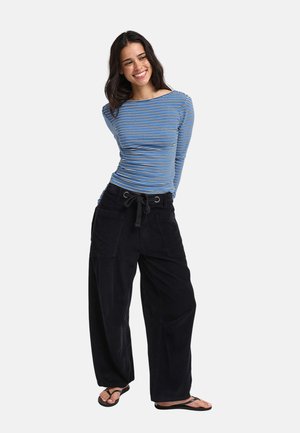 Femme souriante debout, les mains dans le dos, portant un haut à manches longues à rayures bleues, un pantalon large noir et des tongs noires.