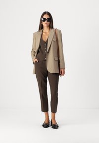Blazer beige ajusté sur un gilet marron avec des boutons et un pantalon ajusté assorti. Lunettes de soleil noires et chaussures en cuir laqué avec des accents.