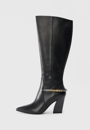 Black leather knee-high boot with a pointed toe and block heel. Features a chain accent in gold and silver tones around the ankle.