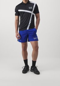 Černé polo tričko s bílými detaily a logem EA7, doplněné modrými šortkami s logem, obojí vyrobené z lehkých materiálů. Černé boty a ponožky.
