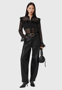 Schwarze transparente Bluse mit Rüschenausschnitt, Knopfverschluss und Spitzelementen; kombiniert mit hochgeschnittenen schwarzen Lederhosen und einer kleinen Handtasche.