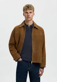 Smeđi suđeni sako s ovratnikom i patentnim zatvaračem, nosi se preko mornarsko plave polo majice. Model stoji s rukama u džepovima.