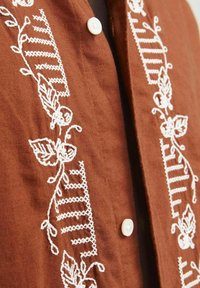 Chemise en lin marron ornée de broderies florales blanches et d'un motif rayé vertical. Détails avec un bouton blanc au col.