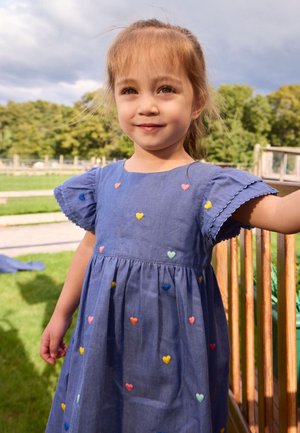 Noor tüdruk helepruuni juustega sinises kleidis, millel on mitmevärvilised tikitud südamed, seisab väljas puidust aiapiirde lähedal.