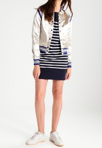 Donna che indossa un vestito a righe blu e bianche, una giacca bomber argentata con polsini blu e sneakers bianche, in piedi contro uno sfondo semplice.
