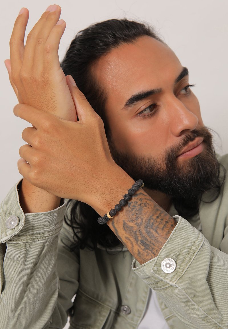 Schwarzes Lavasteinarmband mit runden Perlen und goldenen Akzenten, getragen an einem Handgelenk mit komplizierten Tattoos, vor einem neutralen Hintergrund.