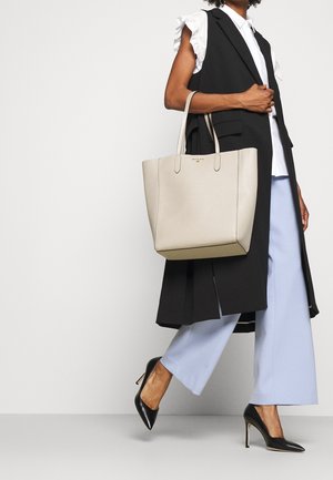 Kobieta w czarnym bezrękawniku, białej bluzce z falbankami, jasnoniebieskich spodniach o szerokich nogawkach, czarnych szpilkach, niosąca dużą beżową torbę typu tote.