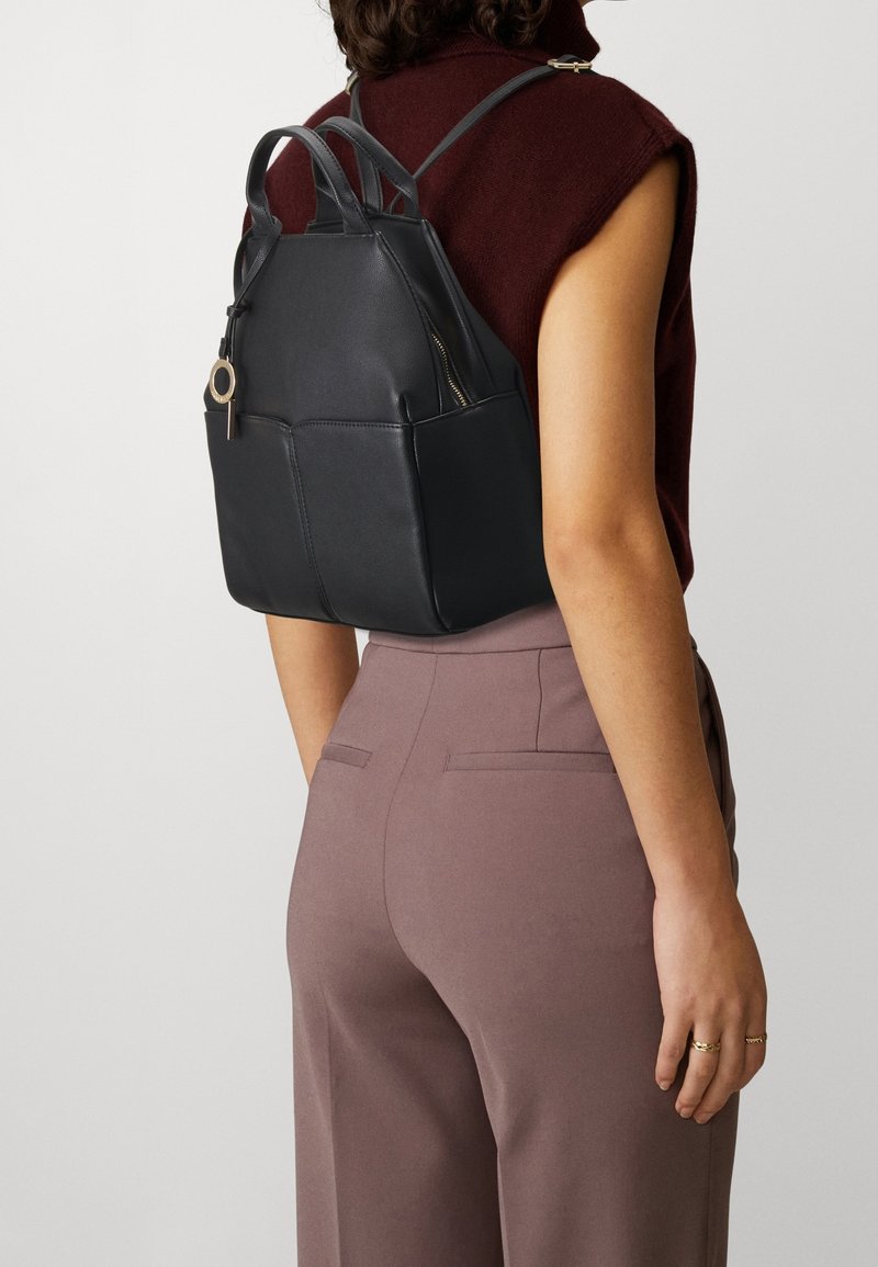 Femme portant un haut sans manches bordeaux et un pantalon mauve, portant un sac à dos en cuir noir avec un porte-clés rond sur la fermeture éclair.