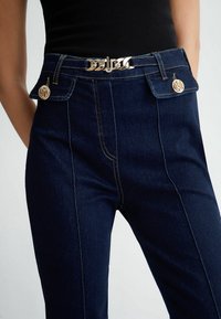 Tmavě modré džínové kalhoty s vysokým pasem, zdobené zlatými kovovými prvky, předními kapsami a kontrastním prošíváním.