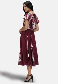 Abito bordeaux con maniche a farfalla corte, motivi astratti bianchi e neri, gonna fluida e design con apertura a chiave sulla schiena. Tacchi neri con cinturini.