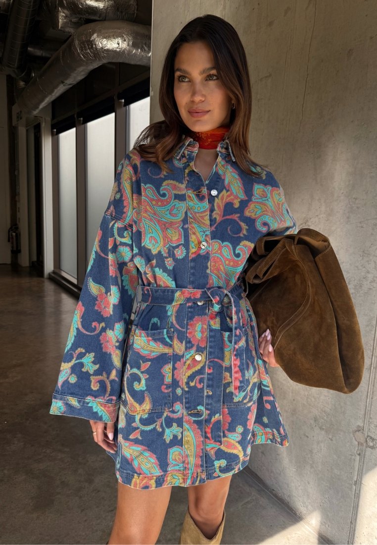 Γυναίκα που φοράει μπλε τζιν φόρεμα με λουλουδάτο μοτίβο και ζώνη, καφέ σουέτ τσάντα και κόκκινο φουλάρι λαιμού σε έναν βιομηχανικό διάδρομο.