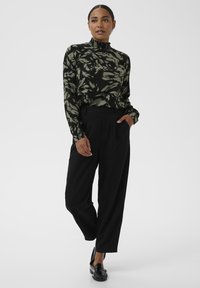 Zwart, langärmelig topje met groen bladpatroon en hoge gerimpelde kraag; gecombineerd met loszittende zwarte broek en zwarte loafers.