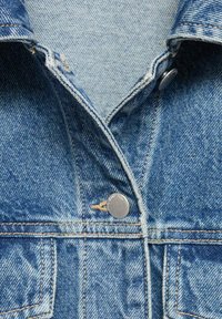 Giacca in denim blu chiaro con chiusura a bottoni, dettagli di cucitura visibili e design del colletto classico. La texture appare leggermente usurata.