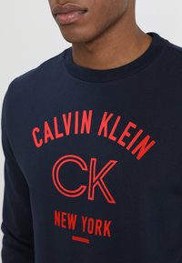 Sudadera de cuello redondo en color azul marino, hecha de algodón, con un texto rojo en negrita que dice "CALVIN KLEIN CK NEW YORK," y con una textura suave.