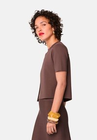 Bruine T-shirt met korte mouwen, een relaxed fit en een ronde halslijn, gecombineerd met gelaagde armbanden in verschillende tinten geel en helder materiaal.