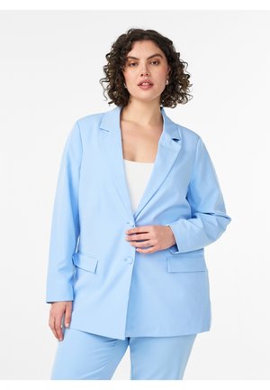 Frau mit lockigem Haar, die einen hellblauen Blazer und passende Hose trägt, posiert mit einer Hand in der Nähe der Blazer-Knöpfe vor weißem Hintergrund.