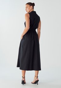 Juodas bepiešis suknelė su apykakle, smarkiai susiaurėjusi juosmens, ir šoninėmis kišenėmis. Ilga, laisvai krintanti sijonas su lygiu paviršiumi; dėvima su juodomis basutėmis.