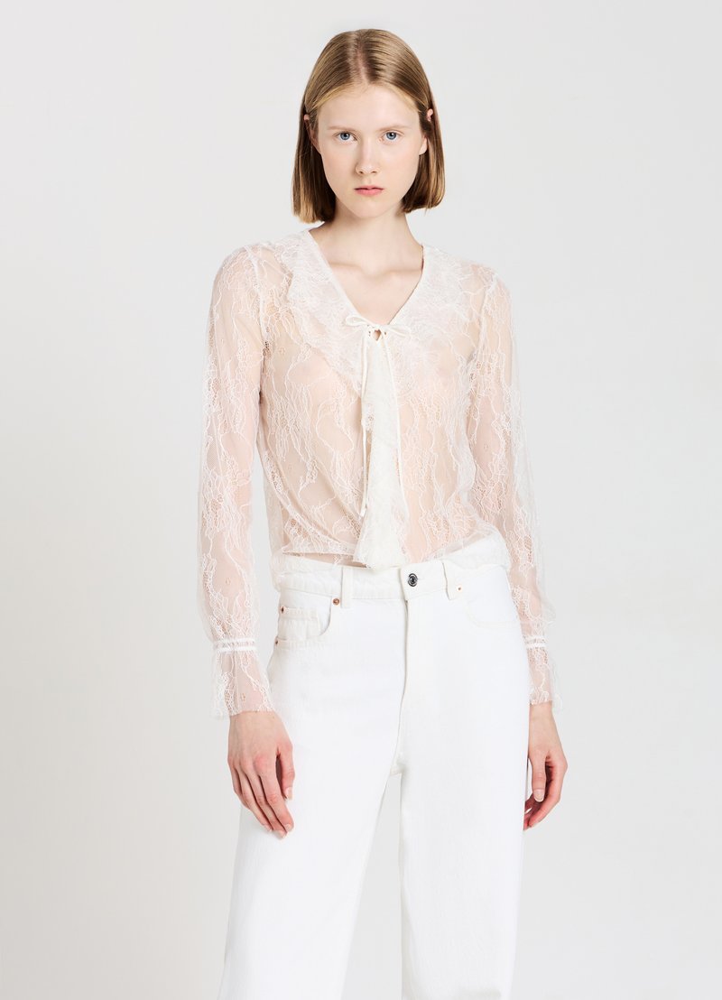 Blusa in pizzo trasparente color pesco, con maniche lunghe, scollo a V e chiusura anteriore con fiocco; abbinata a jeans bianchi a vita alta.