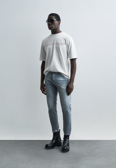 Νεαρός άνδρας που φοράει γυαλιά ηλίου, λευκό φαρδύ t-shirt, ανοιχτόχρωμο τζιν με φθορές και μαύρα δερμάτινα μποτάκια στέκεται μπροστά σε απλό ανοιχτό τοίχο.