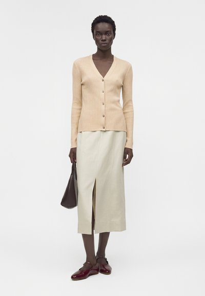 Γυναίκα φοράει μπεζ ριμπ κασμίρ, κρεμ midi φούστα με εμπρός σκίσιμο, μπορντό μπαλαρίνες, κρατώντας σκούρα καφέ τσάντα, στέκεται σε άσπρο φόντο.