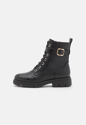 Botas de tobillo de cuero negro con ojales dorados, diseño con cordones, correa texturizada con hebilla, suela gruesa y asa para facilitar el uso.
