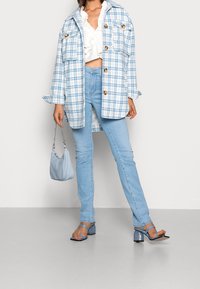 Veste oversize à carreaux bleus avec des boutons, top blanc à volants, jeans skinny bleu clair et talons à lanières assortis. Petit sac bleu clair.