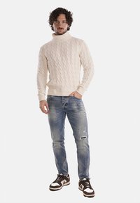 Maglione a trecce color crema con collo alto, abbinato a jeans chiari strappati e sneakers marroni con accenti bianchi.