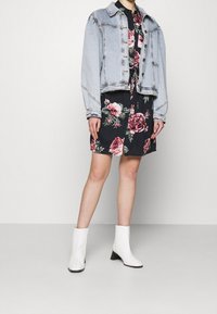 Giacca di jeans azzurro chiaro sopra un vestito a stampa floreale in colori scuri, abbinata a stivali alla caviglia bianchi con tacco marrone.