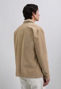 Hombre con cabello oscuro que lleva una chaqueta de pana beige y pantalones blancos, de pie y de perfil contra un fondo claro y liso.