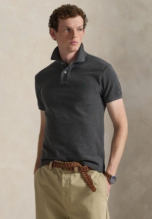 Jeune homme aux cheveux bouclés portant un polo gris foncé, un pantalon beige, une ceinture tressée marron et une montre-bracelet, debout, les mains dans les poches.