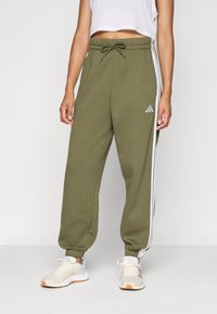 Pessoa vestindo calças de jogging Adidas verde-azeitona com riscas brancas, tornozelos elásticos, cintura com cordão, ténis brancos e uma blusa curta sem mangas branca.