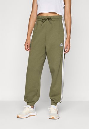 Person, der olivgrüne Adidas-Jogginghosen mit weißen Streifen, elastischen Bündchen, einer Kordelzug-Taille, weißen Sneakers und einem kurzen, weißen ärmellosen Oberteil trägt.