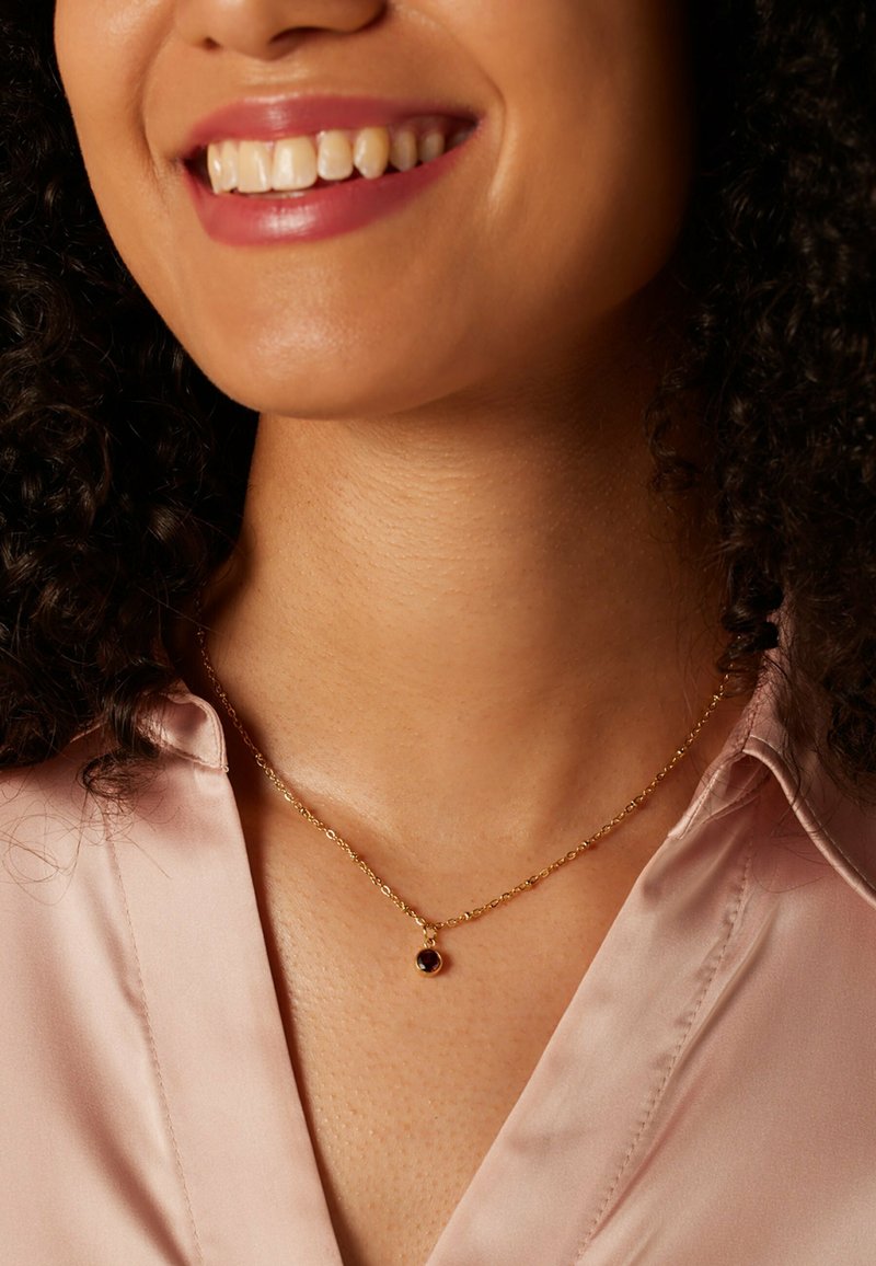 Goldene Kettenhalskette mit einem kleinen, runden Anhänger, der einen dunklen Edelstein zeigt. Das Modell trägt eine blassrosa Bluse. Die glatte Hautstruktur ist sichtbar.