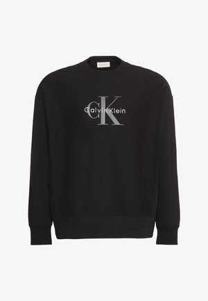 Črna majica z dolgimi rokavi in okroglim izrezom, z logotipom "ck Calvin Klein" v sivi in beli barvi, ki je postavljen na sredino prsnega koša.