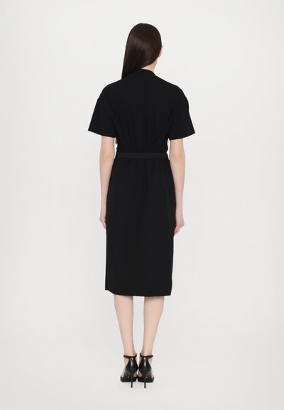 Schwarzes Kurzarmkleid mit einem taillierten Gürtel, knielangem Saum und klarer Silhouette. Kombiniert mit schwarzen Sandalen mit Absatz.