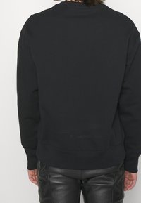 Sweat-shirt noir avec un col rond classique, des poignets et un ourlet côtelés. Le tissu semble doux et peut avoir une légère texture. Porté avec un pantalon en cuir.